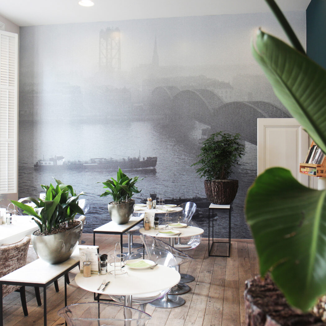 Modern restaurant interieur met witte tafels, transparante stoelen, veel planten en een muurschildering van een stad met brug en rivier.