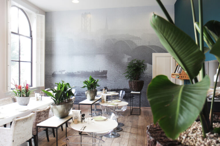Modern restaurant interieur met witte tafels, transparante stoelen, veel planten en een muurschildering van een stad met brug en rivier.