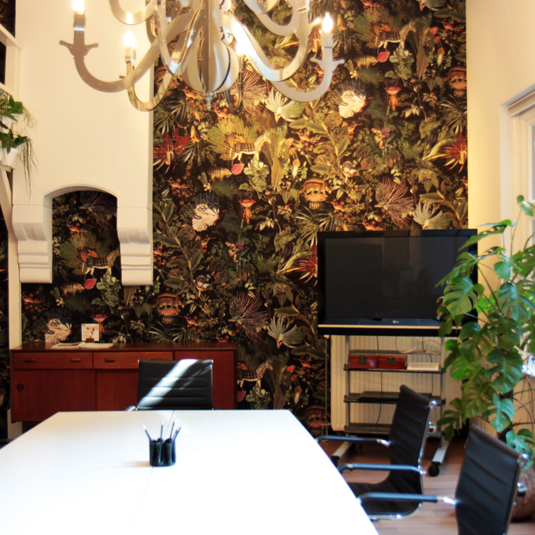 Sfeervolle vergaderruimte met witte tafel, zwarte stoelen, donker jungle behang en diverse planten.