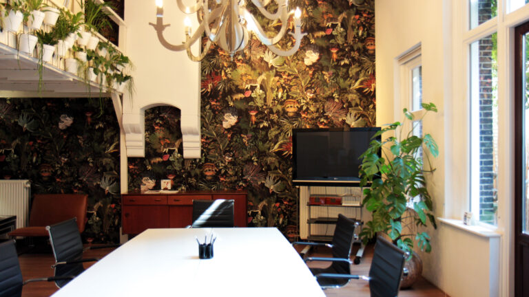 Sfeervolle vergaderruimte met witte tafel, zwarte stoelen, donker jungle behang en diverse planten.