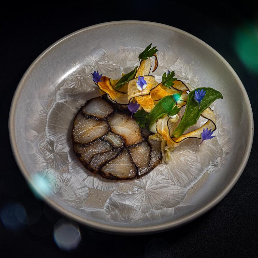 Elegant opgemaakt gerecht met een ronde plak, groentenchips, groene kruiden en paarse bloemen op een grijze keramische schaal.