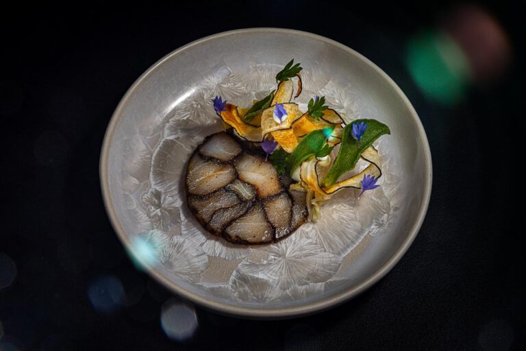 Elegant opgemaakt gerecht met een ronde plak, groentenchips, groene kruiden en paarse bloemen op een grijze keramische schaal.