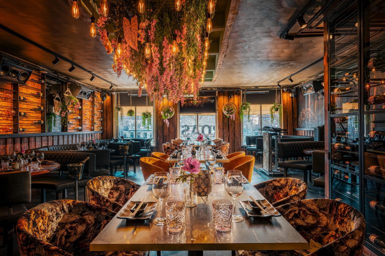 Luxe restaurantinterieur met gedekte tafel, hangende roze bloemen, vintage lampen, houten wijnrekken en ROAST-glazen.