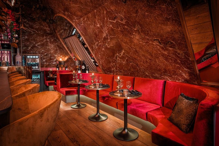 Warm verlichte lounge met rode fluwelen zitbanken, glimmende marmeren muren en bar met oranje krukken.