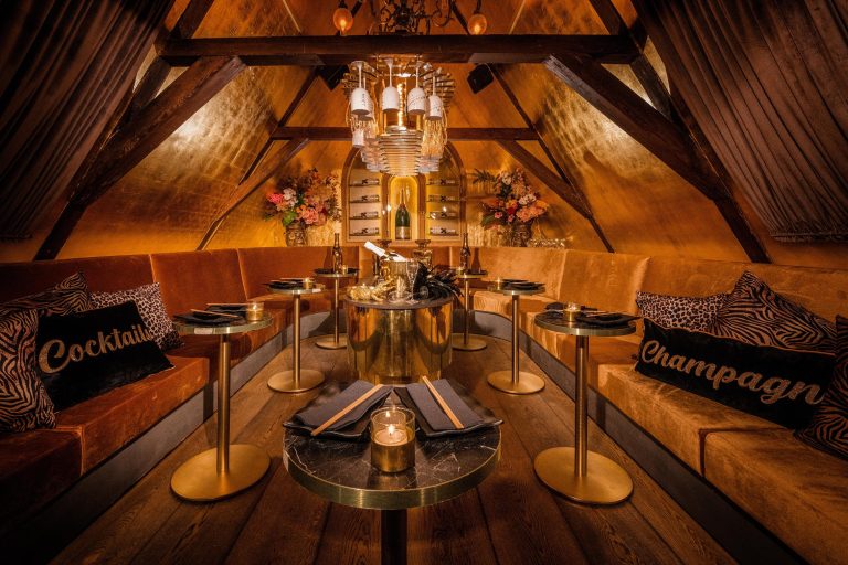 Chic zolderinterieur met oranje fluwelen banken, gouden wanden, champagnekoeler en kussens met Cocktail en Champagne.
