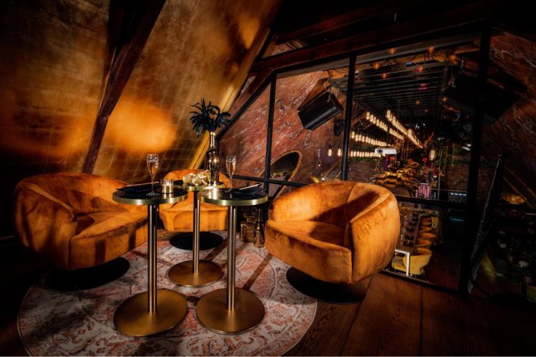 Knusse loungehoek met drie oranje fluwelen fauteuils, gouden tafels met champagne, voor een glazen wand met sfeervolle verlichting.