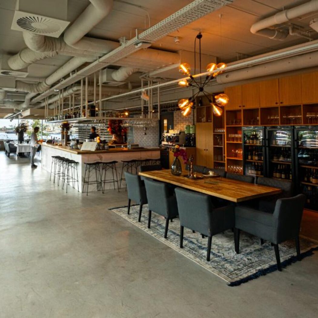Ruim, modern restaurant met houten bar, lange eettafel en een wand vol wijnen.