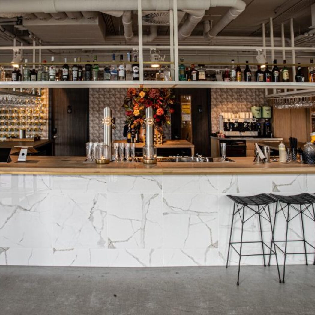 Een sfeervolle, moderne bar met een houten balie, marmeren tegels, wijnrekken, biertappen en zwarte barkrukken.