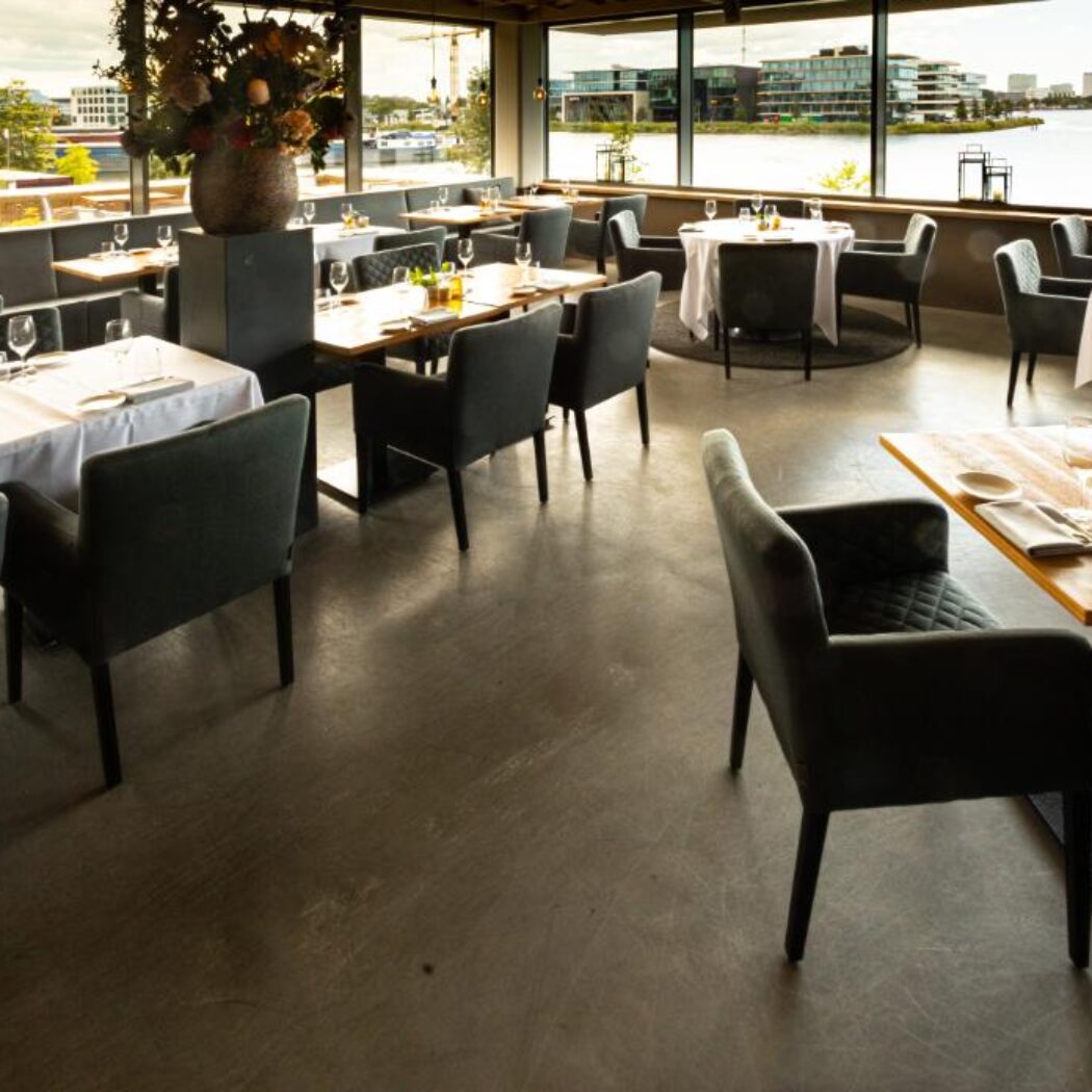 Ruim restaurantinterieur met moderne gedekte tafels, donkere stoelen en een panoramisch uitzicht over het water.