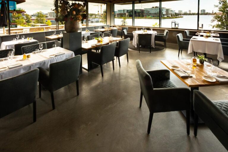 Ruim restaurantinterieur met moderne gedekte tafels, donkere stoelen en een panoramisch uitzicht over het water.