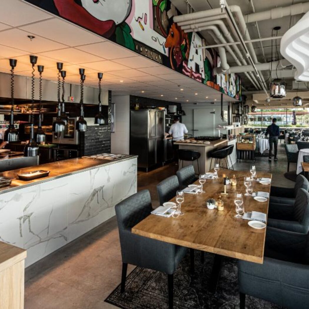 Stijlvol restaurantinterieur met open keuken, marmeren bar, houten tafels en een kleurrijke muurschildering.