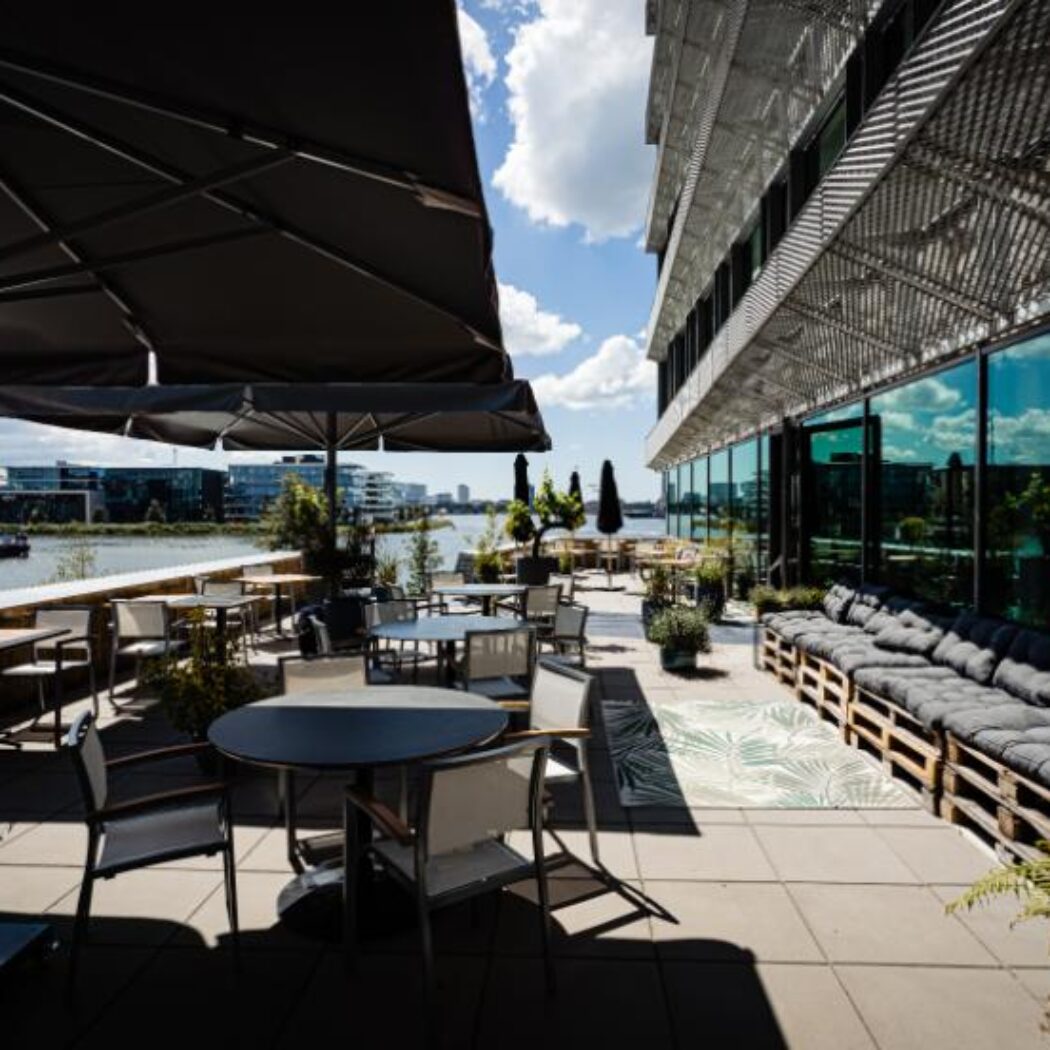 Uitgestrekt zonnig terras met diverse tafels, stoelen en loungebanken, grote parasols en uitzicht op water en moderne gebouwen.
