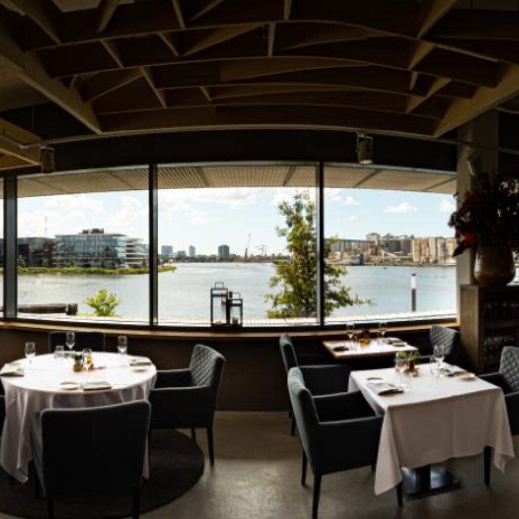 Modern restaurant met gedekte tafels, donkere stoelen en breed uitzicht op de rivier, stad en een COSCO schip.