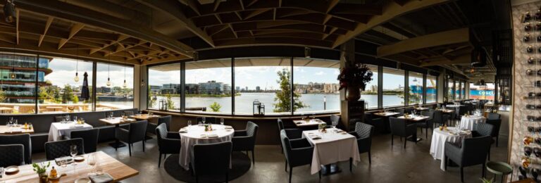 Modern restaurant met gedekte tafels, donkere stoelen en breed uitzicht op de rivier, stad en een COSCO schip.