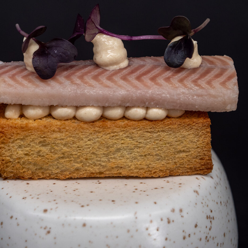 Roze vis met chevronpatroon, romige crème en paarse microgreens op krokante toast, geserveerd op een gespikkeld bord.