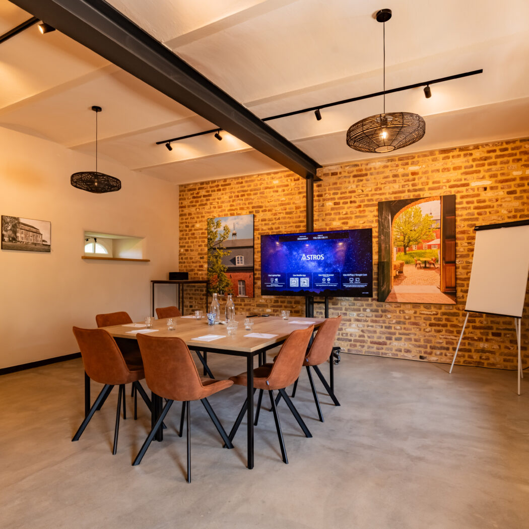 Moderne vergaderruimte met houten tafel, oranjebruine stoelen, groot scherm met ASTROS en bakstenen muur.