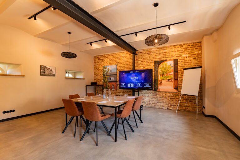 Moderne vergaderruimte met houten tafel, oranjebruine stoelen, groot scherm met ASTROS en bakstenen muur.