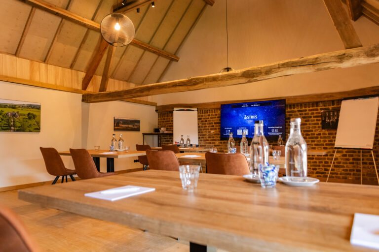 Rustieke vergaderzaal met houten balken, tafels met water, bruine stoelen en een presentatiescherm met ASTROS.