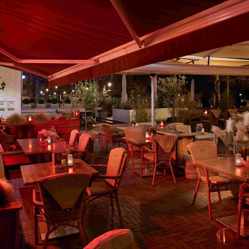 Gezellig verlicht restaurantterras s avonds met rode luifels, houten tafels en comfortabele zitjes.