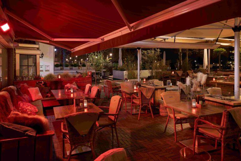 Gezellig verlicht restaurantterras s avonds met rode luifels, houten tafels en comfortabele zitjes.