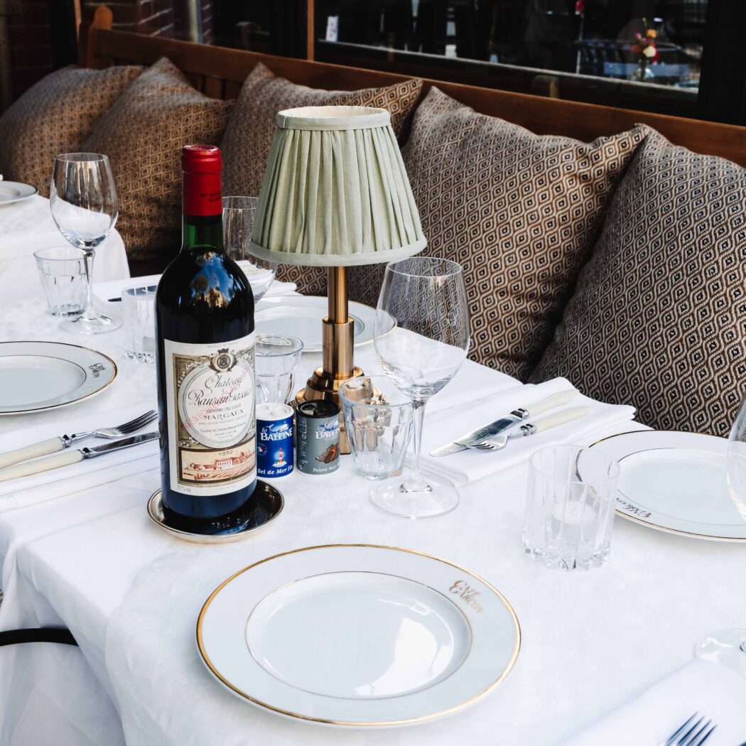 Een gedekte restauranttafel buiten met Château Margaux rode wijn, borden, glazen en een tafellamp met geplooide kap.