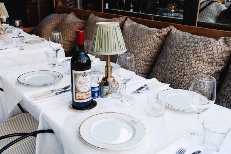 Een gedekte restauranttafel buiten met Château Margaux rode wijn, borden, glazen en een tafellamp met geplooide kap.