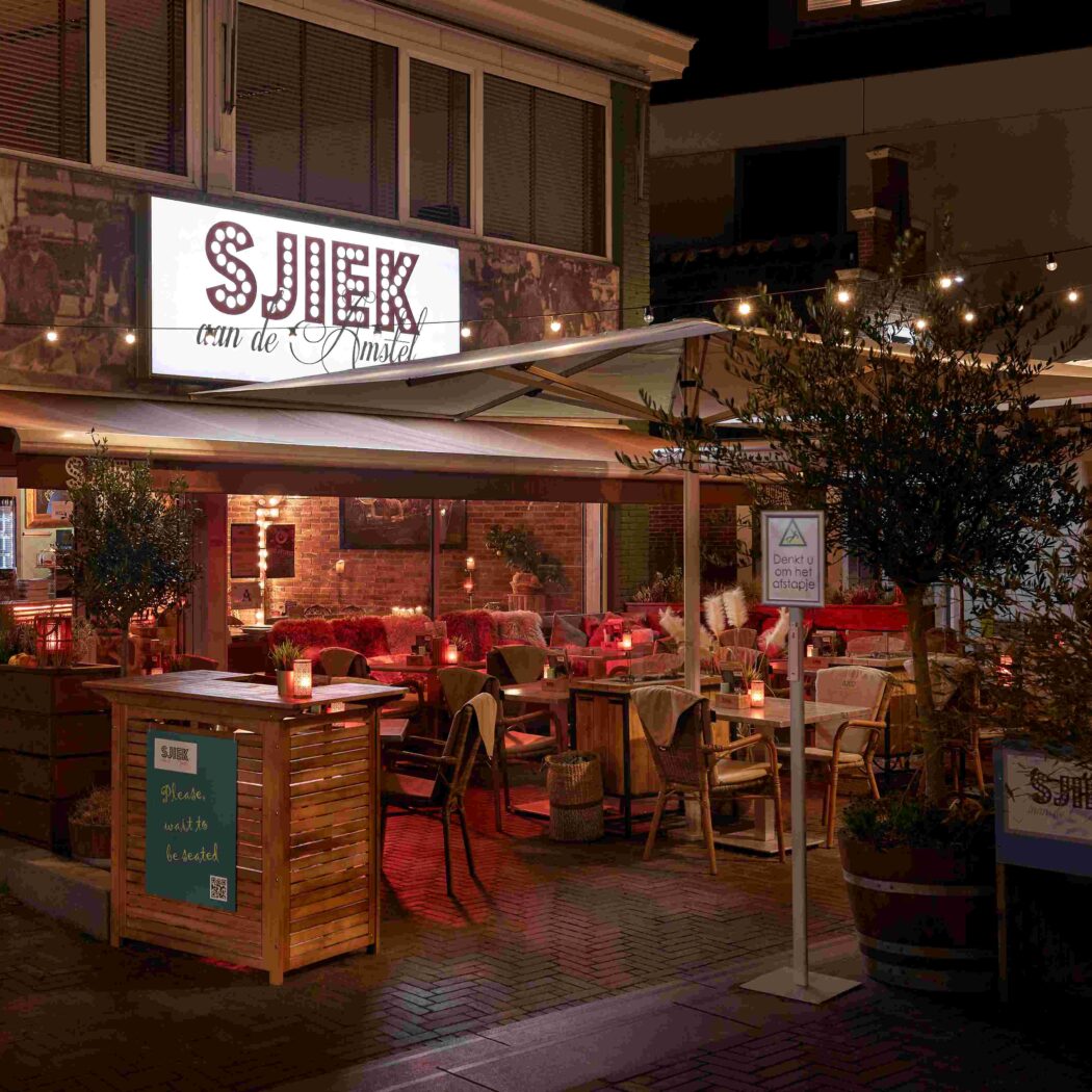 Sfeervol verlicht avondterras van restaurant SJIEK van de Amstel met kaarsen en een Please wait to be seated bord.