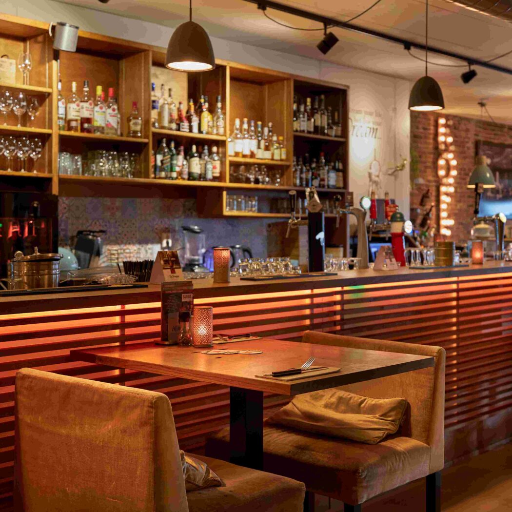 Sfeervolle restaurantbar met Chouffe tap, gevulde flessenrekken, een oranje verlichte houten toonbank en fluwelen stoelen.