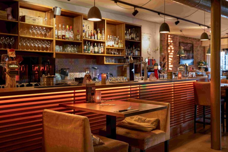 Sfeervolle restaurantbar met Chouffe tap, gevulde flessenrekken, een oranje verlichte houten toonbank en fluwelen stoelen.