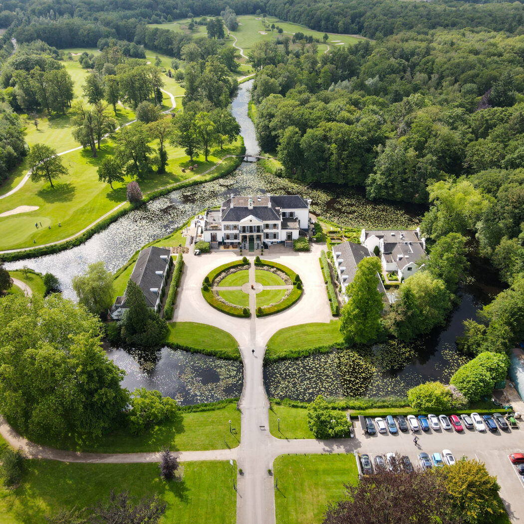 Luchtfoto van een statig landgoed met wit herenhuis, omringd door water, golfbanen en groene bossen.