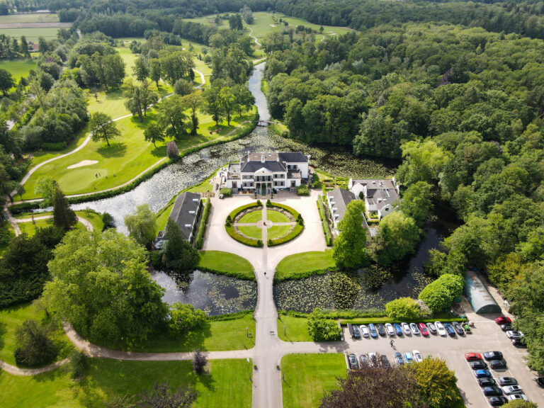 Luchtfoto van een statig landgoed met wit herenhuis, omringd door water, golfbanen en groene bossen.