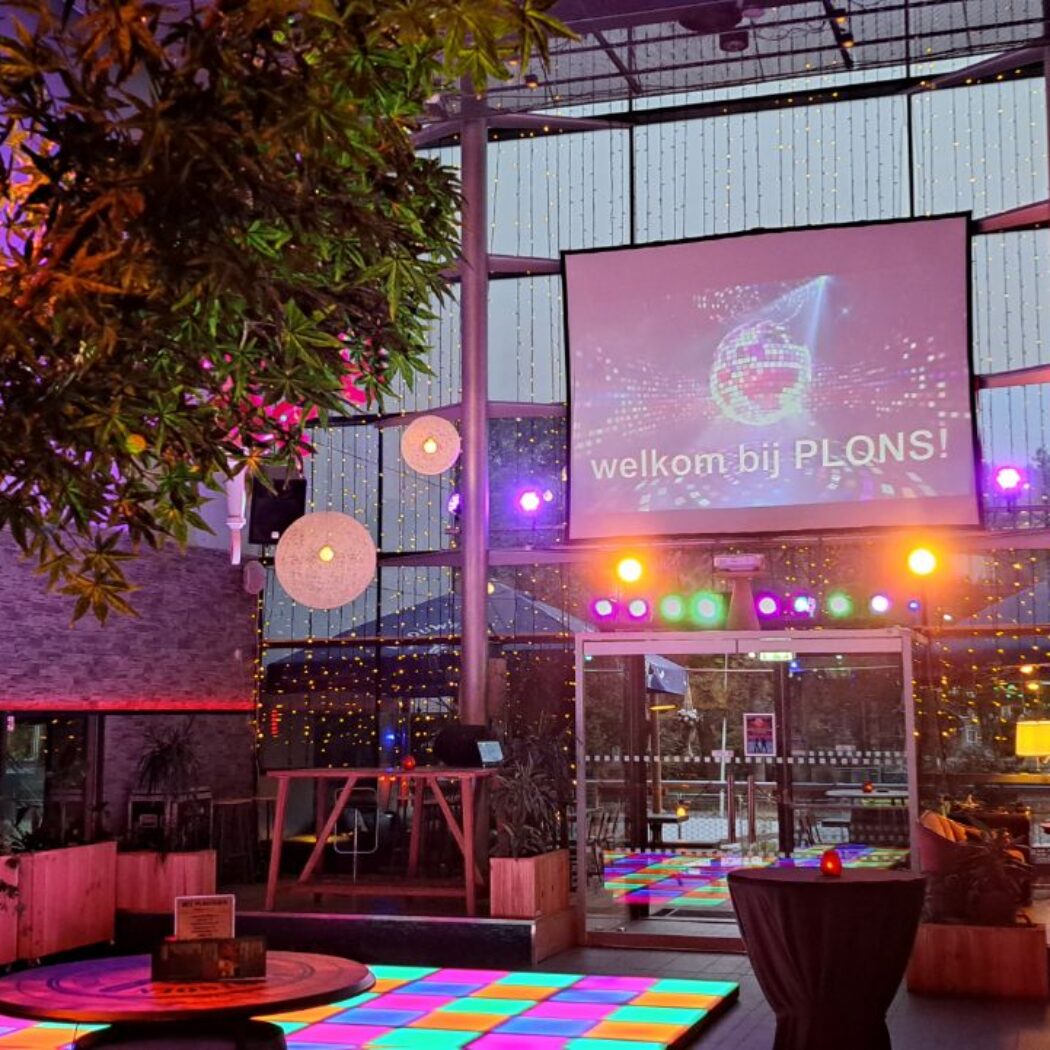 Levendige feestlocatie met kleurrijke dansvloer, scherm met welkom bij PLQNS! en een verlichte boom.