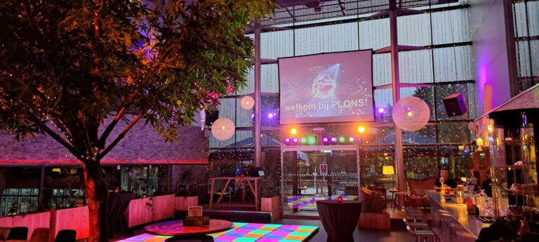 Levendige feestlocatie met kleurrijke dansvloer, scherm met welkom bij PLQNS! en een verlichte boom.