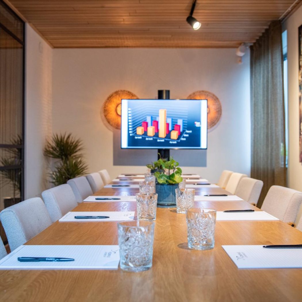 Moderne vergaderzaal met een lange houten tafel, stoelen, tv met staafdiagram en kleurrijke kunst aan de muur.