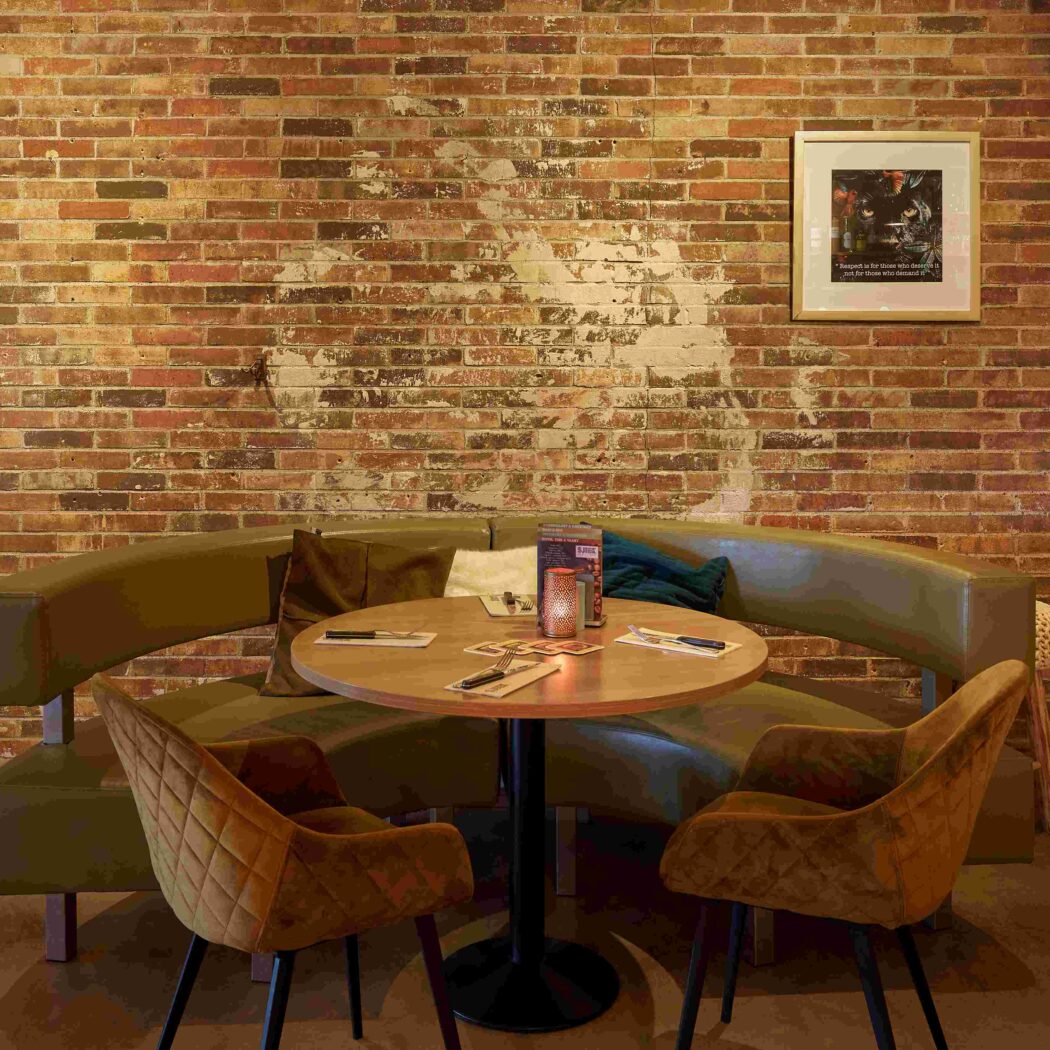 Sfeervolle zithoek met olijfgroene bank, ronde houten tafel en twee fluwelen stoelen voor een bakstenen muur.