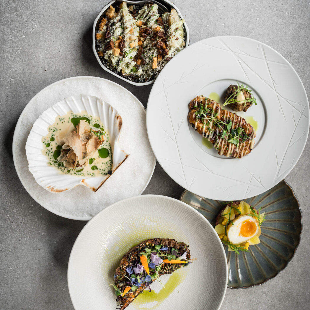 Vijf culinaire gerechten op diverse designborden: salade, vis in schelp, gegrilde filet, plantaardig gerecht en ei-voorgerecht.