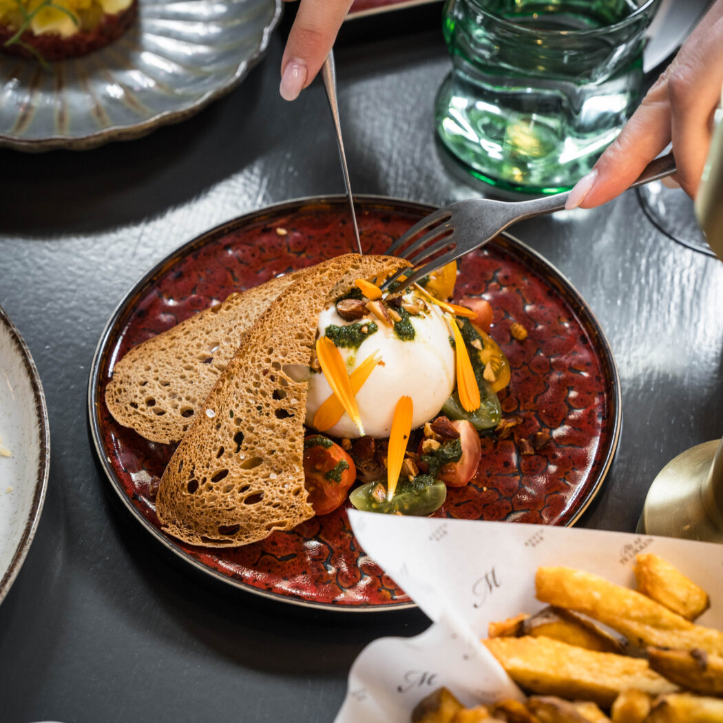 Handen snijden burrata met toast, cherrytomaten, pesto en oranje bloemblaadjes op een rode schaal.