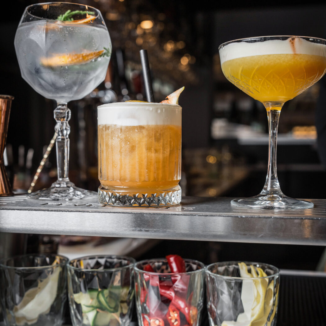 Drie diverse cocktails met schuim en fruit op een barplank, met glazen vol komkommer, pepers en citrusgarnituren eronder.