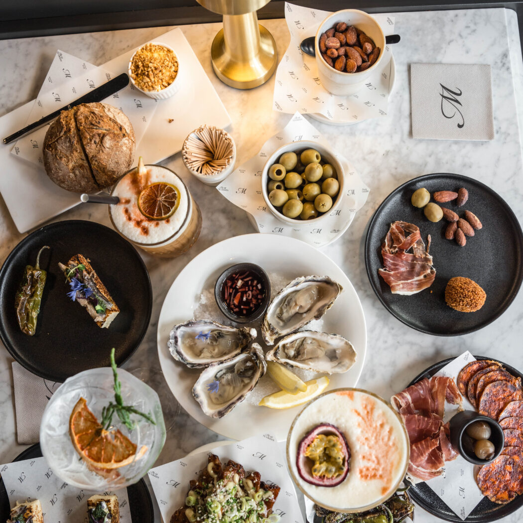 Rijkelijk gedekte marmeren tafel met verse oesters, Spaanse charcuterie, diverse hapjes en kleurrijke cocktails.