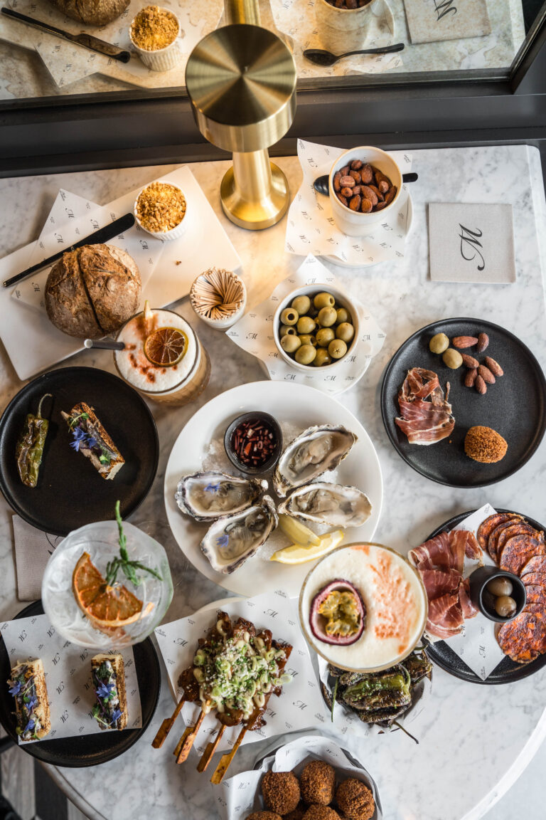 Rijkelijk gedekte marmeren tafel met verse oesters, Spaanse charcuterie, diverse hapjes en kleurrijke cocktails.
