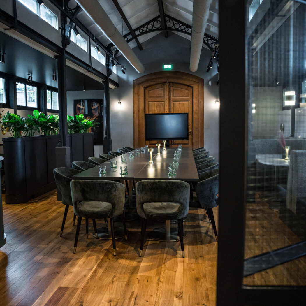 Moderne vergaderzaal met lange tafel, fluwelen stoelen, tv, eikenhouten vloer en planten, onder een hoog industrieel plafond.