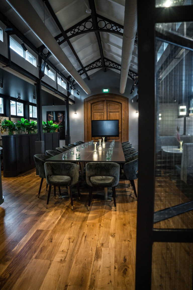 Moderne vergaderzaal met lange tafel, fluwelen stoelen, tv, eikenhouten vloer en planten, onder een hoog industrieel plafond.