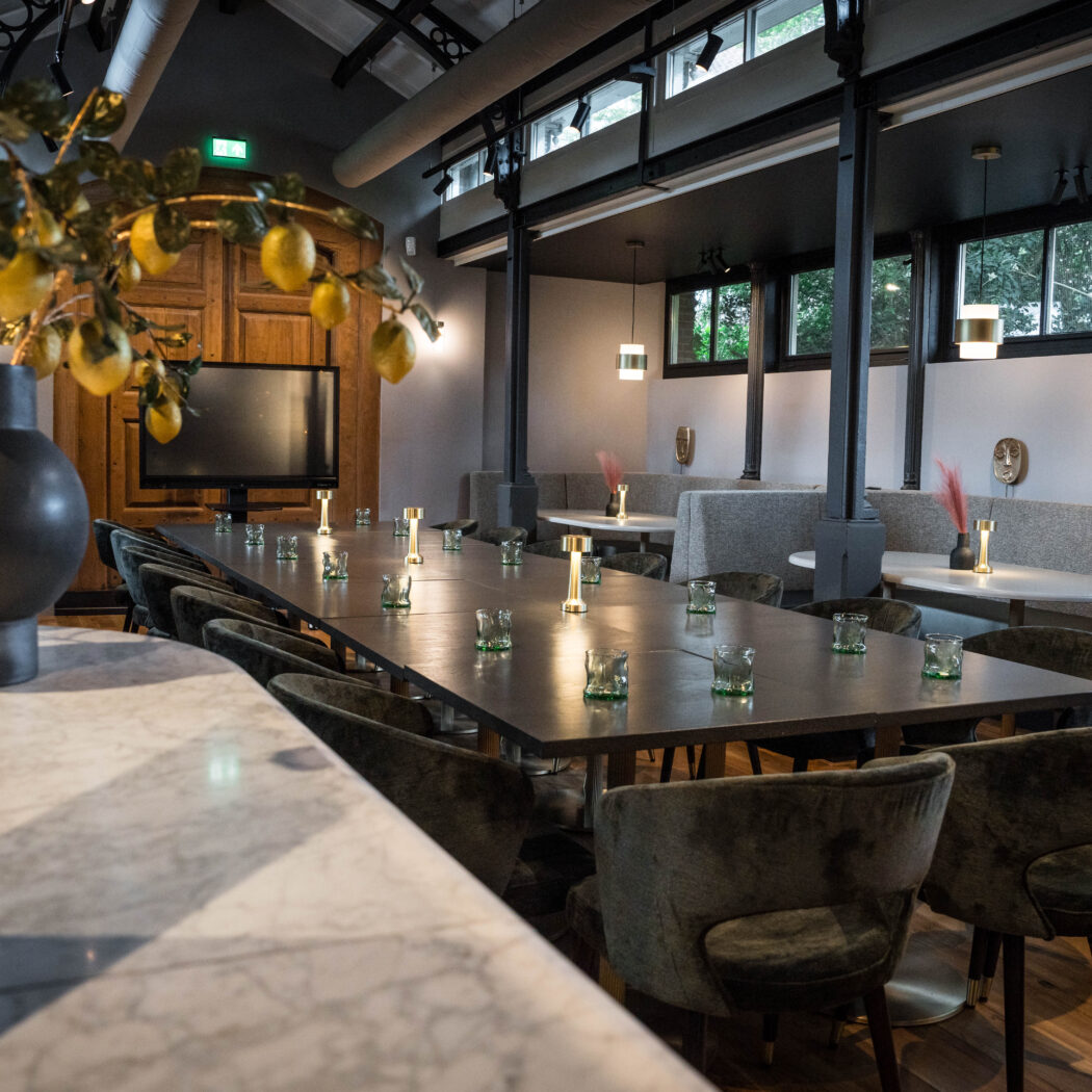 Moderne evenementenruimte met lange tafel, donkergroene stoelen, sfeervolle verlichting en raamzitjes.