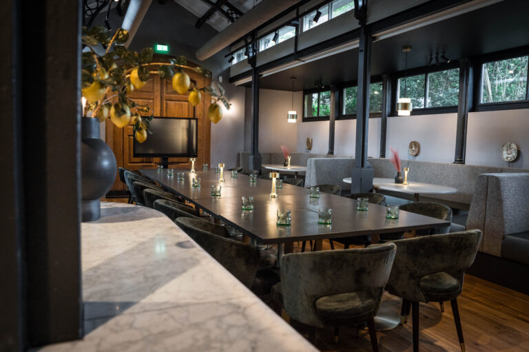 Moderne evenementenruimte met lange tafel, donkergroene stoelen, sfeervolle verlichting en raamzitjes.