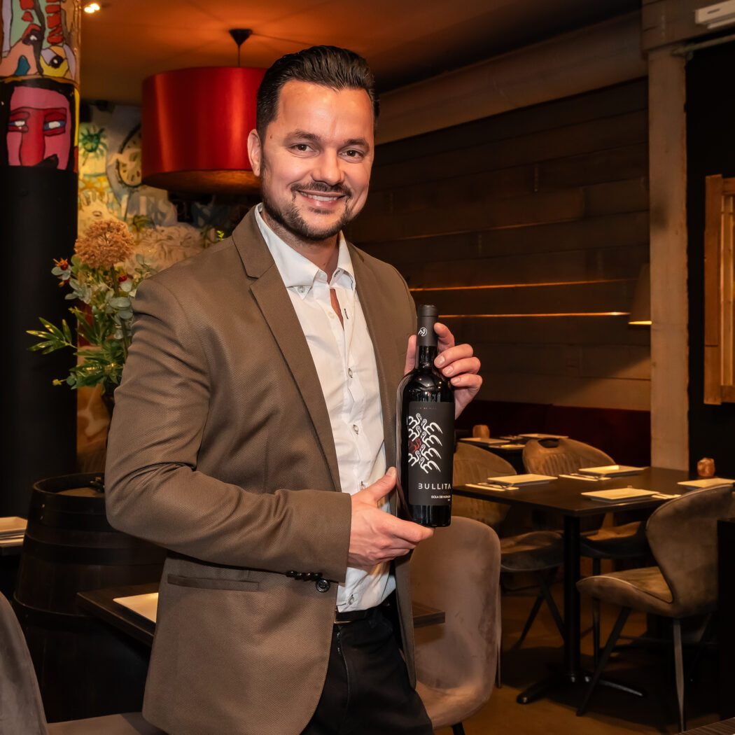 Glimlachende man in bruin pak presenteert een zwarte BULLITA wijnfles met opvallend label in een sfeervol restaurant.