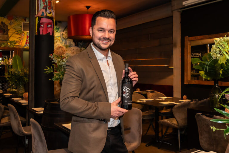 Glimlachende man in bruin pak presenteert een zwarte BULLITA wijnfles met opvallend label in een sfeervol restaurant.