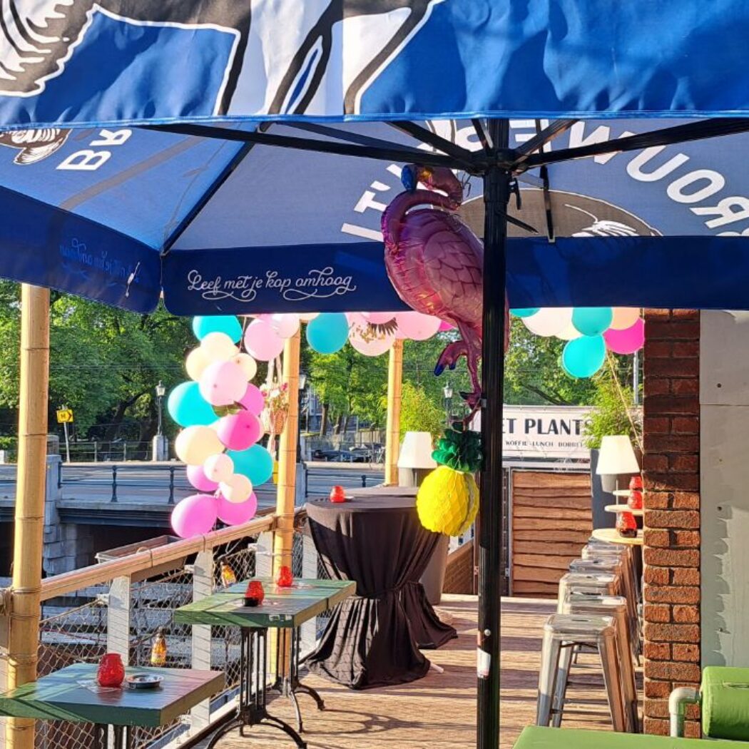 Feestelijk terras onder blauwe parasol met Leef met je kop omhoog tekst, roze flamingo en kleurrijke ballonnen, aan een gracht.
