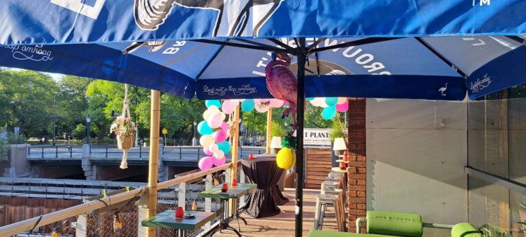 Feestelijk terras onder blauwe parasol met Leef met je kop omhoog tekst, roze flamingo en kleurrijke ballonnen, aan een gracht.