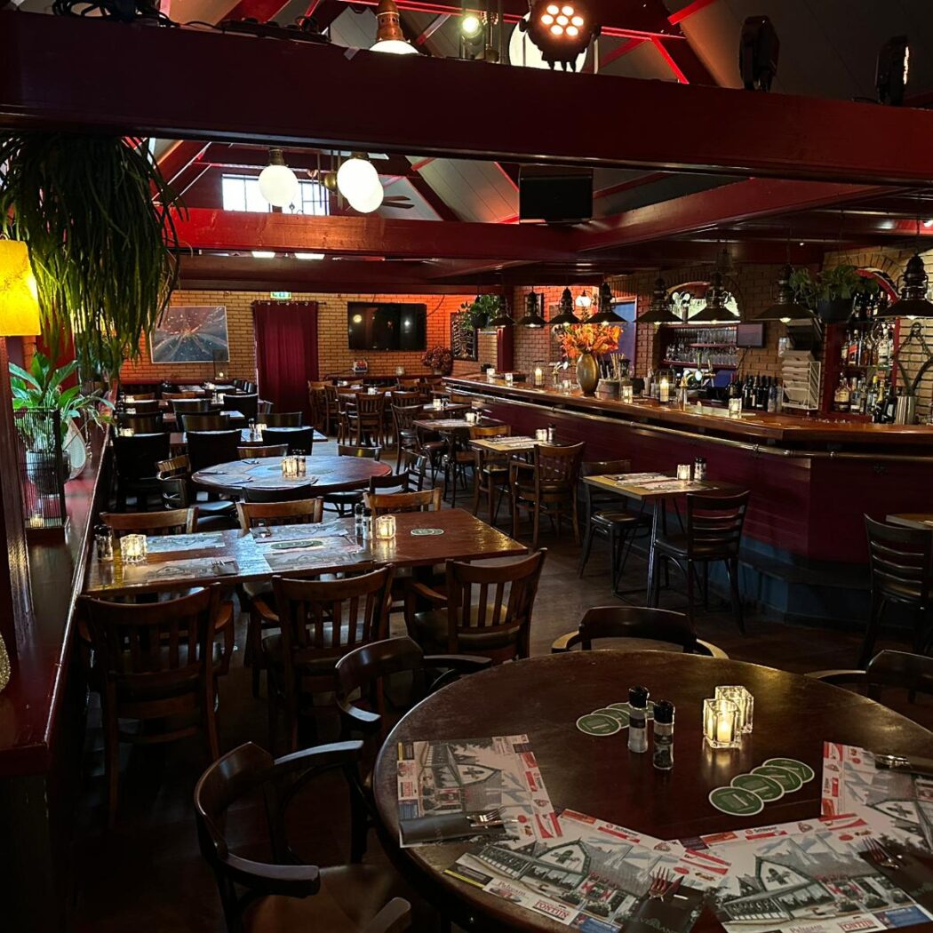 Gezellig restaurantinterieur met houten tafels, rode balken, een bar en placemats met Pelgrim Poortje, verlicht door kaarslicht.