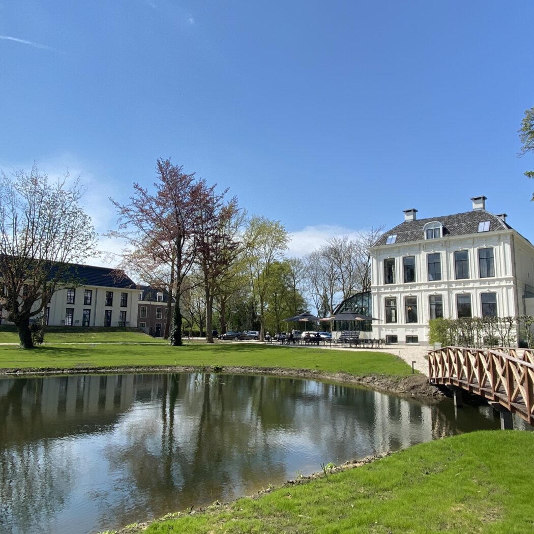 Stijlvol wit landhuis met terras, weerspiegeld in een vijver met houten brug op een zonnige dag.